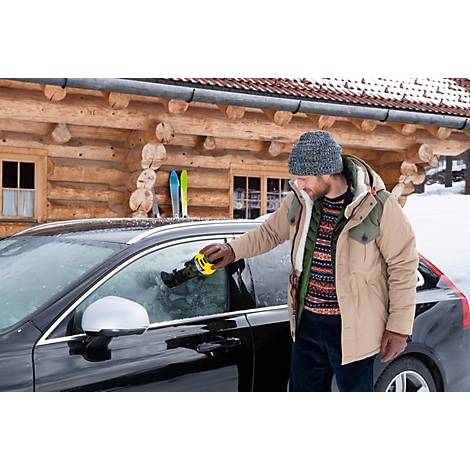Un homme gratte la glace sur le pare-brise d'une voiture en hiver. En arrière-plan, une maison en bois avec des skis.
