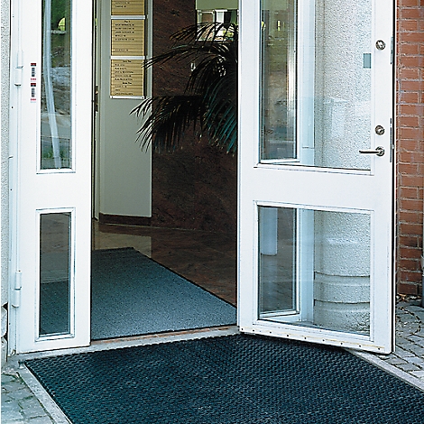 Offene weiße Eingangstür mit Glastüren. Schwarze Türmatte davor. Dahinter eine dunkelbraune Wand und eine Pflanze.