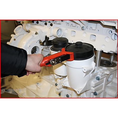 Ein Mechaniker hält einen roten Ölfilterschlüssel. Er arbeitet an einem weißen Motor. Rechts befindet sich ein schwarzer Ölfilter.