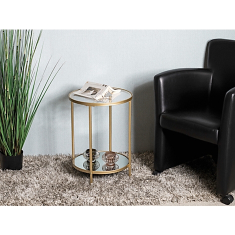 Table d'appoint dorée avec plateaux en verre et décorations. À côté, un fauteuil noir et une plante d'intérieur.