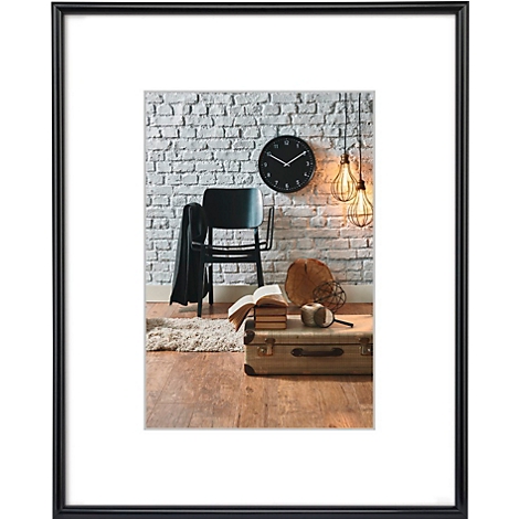 Une photo dans un cadre noir. Elle montre une pièce avec un mur de briques et une horloge. Une chaise, une valise, des livres, des lampes et un tapis sont visibles.