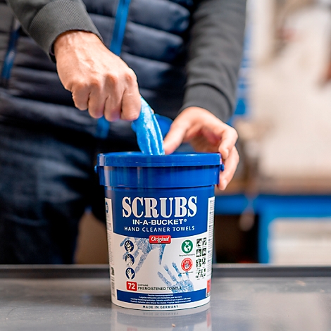 A person is taking a blue cleaning towel out of a blue bucket. The bucket says: SCRUBS IN-A-BUCKET HAND CLEANER TOWELS.