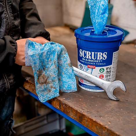 Persoon houdt een gebruikte schoonmaakdoek vast. Daarnaast een emmer schoonmaakdoekjes, een moersleutel op een houten oppervlak.