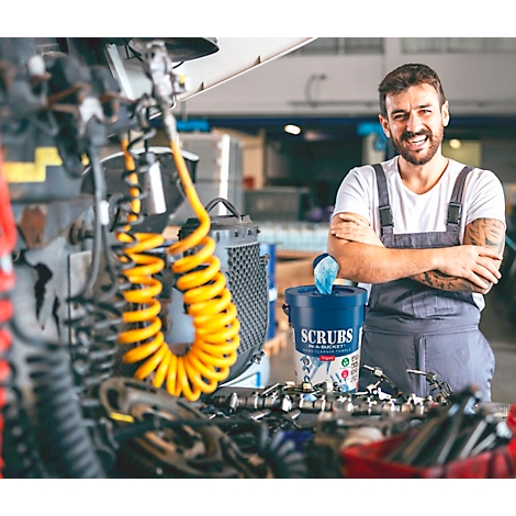 Ein Mechaniker steht in einer Werkstatt vor einem Eimer mit Reinigungstüchern, die Arme verschränkt. Er lächelt in die Kamera.