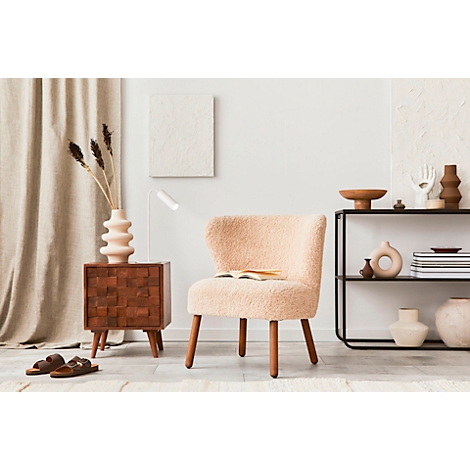 Gezellige woonkamer met crèmekleurige fauteuil, nachtkastje met vaas en boek. Een plank met decoratieve objecten, helder licht.