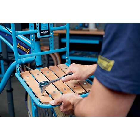 Close-up: Persoon die met een sleutel de houten latten van een bagagedrager van een fiets vastschroeft. Blauwe fiets en gereedschap. Zicht in een werkplaats.