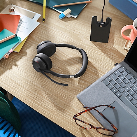 Black headset on a wooden desk, next to a laptop and glasses. In the background notes and a charging station.