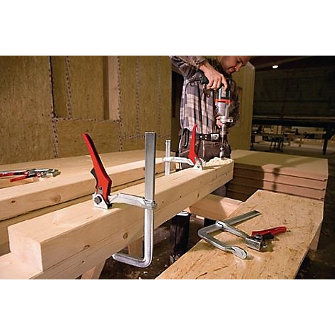Een man boort hout met een boormachine. Klemmen fixeren het hout. Meer houten planken liggen ernaast.