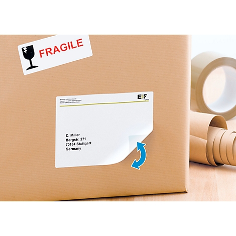 Boîte en carton avec une étiquette d'adresse qui se décolle, à côté d'un rouleau de ruban adhésif. Autocollant avec "Fragile".