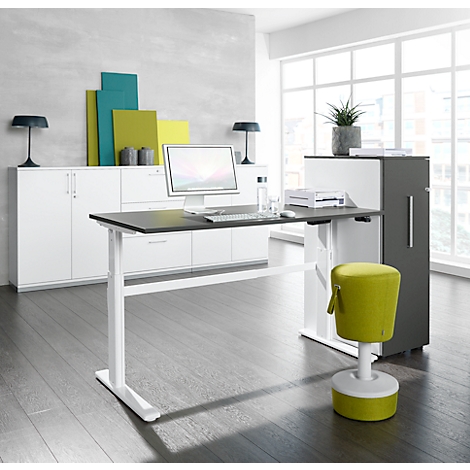 Desk with black top, white frame and green stool in front of a window front. Cabinets in the background.