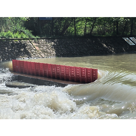 Roter Damm im Fluss. Das Wasser fliesst über eine Betonkonstruktion. Bäume im Hintergrund.