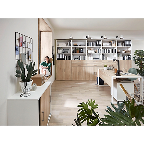 Une femme est assise sur un rebord de fenêtre dans un bureau lumineux. Meubles en bois clair, plantes et design moderne.