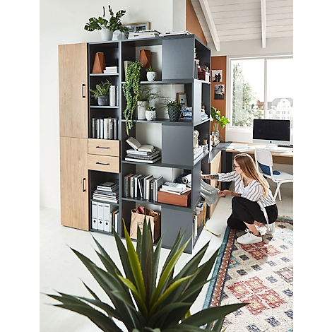 A woman in a modern home office looks at a newspaper next to a large bookcase filled with books and plants.
