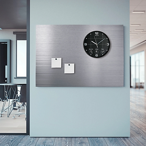 Horloge mural noir avec des post-it sur une plaque métallique sur fond bleu clair. Un couloir de bureau est visible au loin.