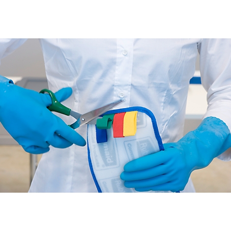 Une personne en blouse blanche et gants bleus coupe des languettes colorées sur un morceau de tissu avec des ciseaux.