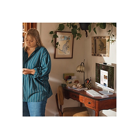 Eine Frau steht in einem Zimmer neben einem Schreibtisch. Sie trägt ein blaues gestreiftes Hemd und Jeans. Auf dem Schreibtisch steht ein All-in-One-Computer.