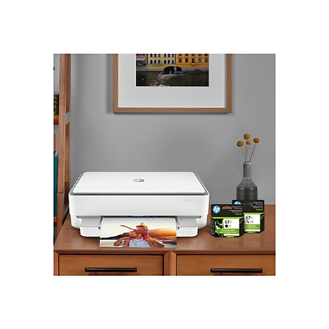 Ein weißer HP Envy 6055-Drucker steht auf einem braunen Holztisch, darunter ein Farbfoto. Rechts stehen Tintenpatronen und eine Vase mit gelben Blüten.