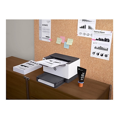 Ein weißer HP-Laserdrucker druckt ein Diagramm; auf einer Kork-Pinnwand hängen Dokumente. Auf dem Tisch steht eine Tonerkartusche.