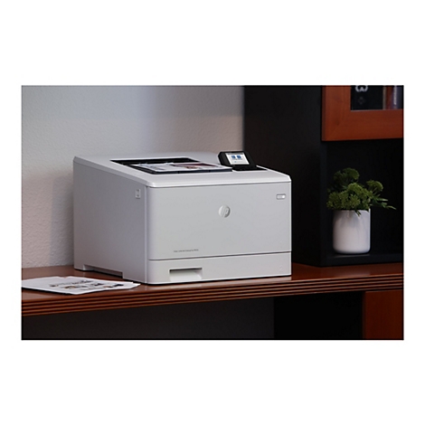 Grauer Drucker auf braunem Schreibtisch. Kleines Display und HP-Logo. Auf dem Tisch liegen Blätter und eine grüne Pflanze.