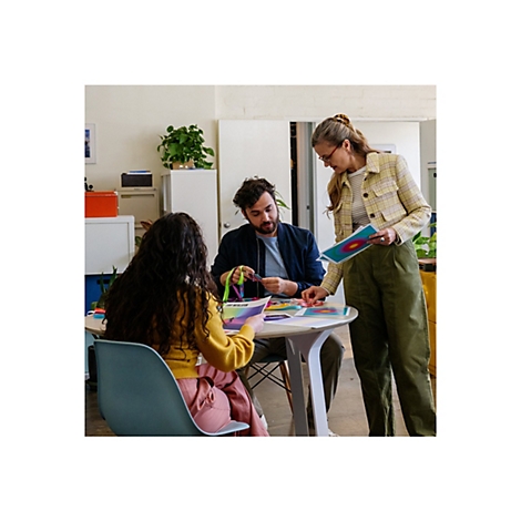 Drei Personen besprechen farbige Designvorlagen an einem runden Tisch. Eine Frau zeigt auf ein Blatt, eine weitere hält ein Blatt, ein Mann hält farbige Bänder.