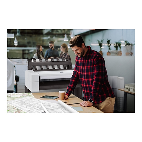 Ein Mann in einem rot-schwarz karierten Hemd, der sich über einen Schreibtisch beugt, auf dem sich Baupläne, ein Tablet und ein Kaffeebecher befinden. Im Hintergrund stehen Personen vor einem Großformatdrucker.