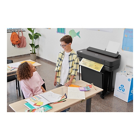 Zwei Schüler stehen an einem Schreibtisch mit Schulmaterialien. Ein schwarzer Drucker druckt ein Dokument. Im Hintergrund eine Wand mit Zeichnungen.