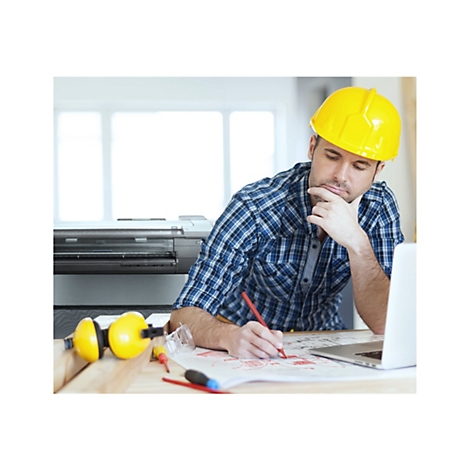 Ein Mann mit gelbem Helm zeichnet auf einem Plan; Laptop, Drucker, Gehörschutz und Brille liegen am Tisch.