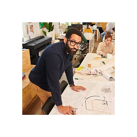 Ein Mann mit Brille beugt sich Ã¼ber BauplÃ¤ne am Tisch, weitere Personen arbeiten im Hintergrund.