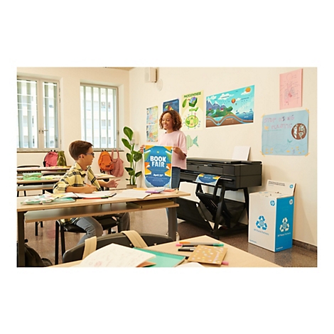 Zwei Schüler in einem Klassenzimmer. Ein Mädchen hält ein Plakat für eine Buchmesse. Auf dem Tisch steht ein Drucker. Auf dem Boden steht eine Recyclingbox.