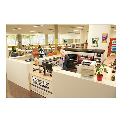 Eine Frau an der Theke des „University Print Center“, befüllt einen Drucker. Im Hintergrund sitzen Leute an Tischen oder in Sitzsäcken.