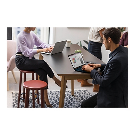 Zwei Personen sitzen an einem Tisch und arbeiten an Laptops. Im Hintergrund stehen weitere Personen.