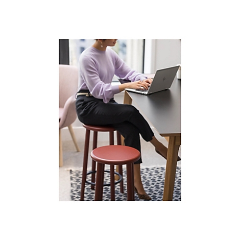 Eine Frau arbeitet an einem Laptop, der auf einem Tisch steht. Sie sitzt auf einem runden Barhocker in einem hellen Raum.