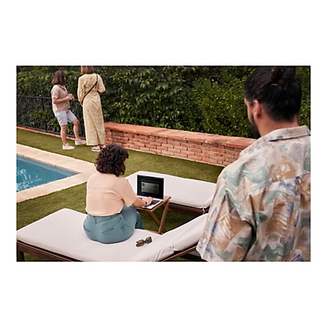 Eine Frau sitzt auf einer Liege und arbeitet mit einem Laptop, im Hintergrund weitere Personen am Pool.