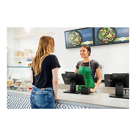 Eine Kundin bestellt an der Theke in einem Restaurant. Die VerkÃ¤uferin lÃ¤chelt und blickt die Kundin an. Hinter ihr sind zwei Bildschirme mit Salatwerbung.