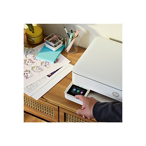 Weißer Drucker auf einem Schreibtisch. Ein Finger berührt den Touchscreen. Auf dem Schreibtisch liegen Papier, Stifte und andere Gegenstände.