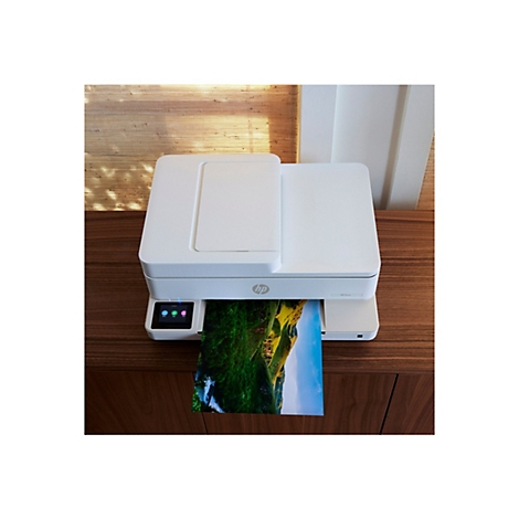 Weißer HP Envy Drucker, druckt ein Farbfoto eines grünen Hügels mit blauem Himmel. Drucker steht auf dunklem Holzschrank.