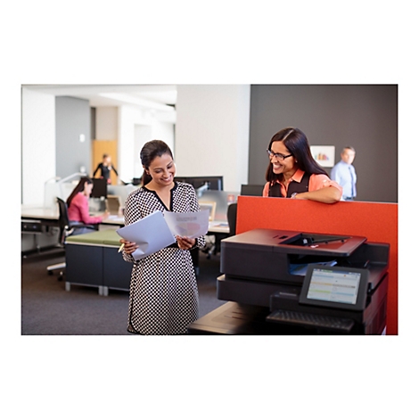 Zwei Frauen im Büro lächeln, eine hält Papiere, die andere lehnt sich an eine rote Trennwand mit Drucker.