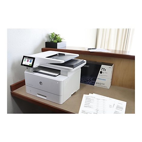 Ein weißer HP Laserdrucker steht auf einem Holzschreibtisch. Auf dem Bedienfeld werden Symbole für Kopieren, Scannen und Drucken angezeigt. Daneben steht eine schwarze Tintenpatrone und Papier.