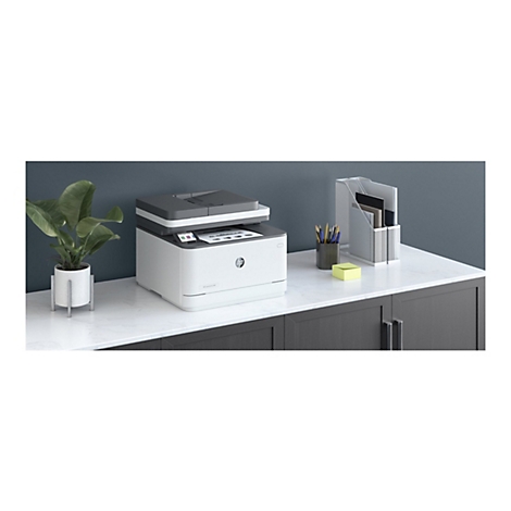 Ein weiß-grauer HP-Laserdrucker steht auf einer weißen Oberfläche. Im Hintergrund sind Büromaterialien und eine Topfpflanze zu sehen.