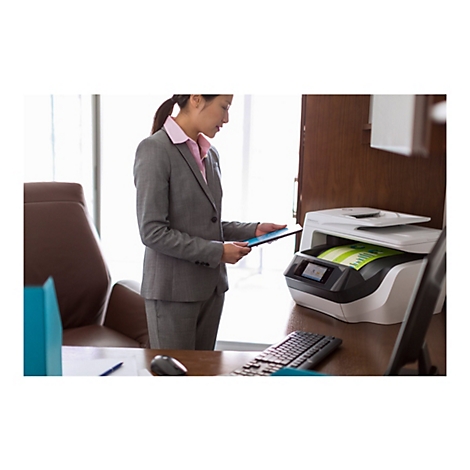Eine Frau in einem grauen Anzug steht an einem Schreibtisch und hält ein Tablet vor einem Drucker. Der Drucker, eine HP OfficeJet Pro 7740, ist geöffnet und druckt ein Dokument.