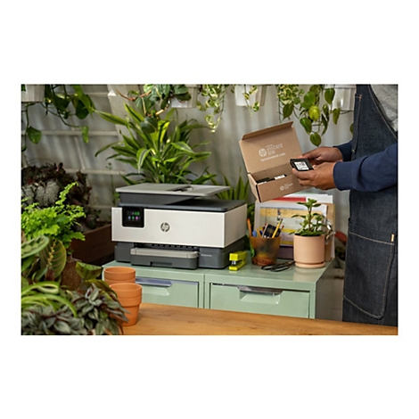 Personne tenant une cartouche d'encre à côté d'une imprimante dans un espace de travail vert. Des plantes entourent l'installation.