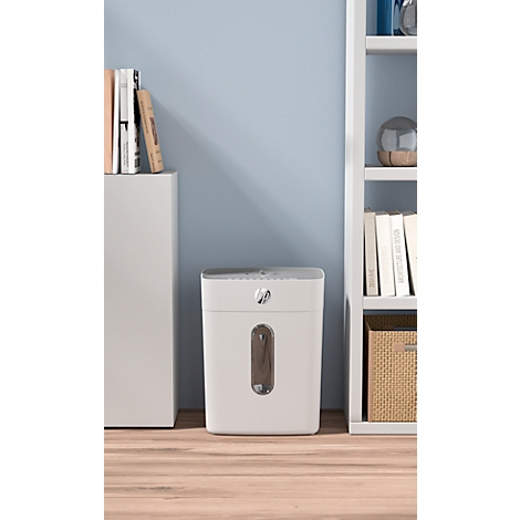 White paper shredder in front of a light blue background. A shelf on the right, a cabinet on the left.