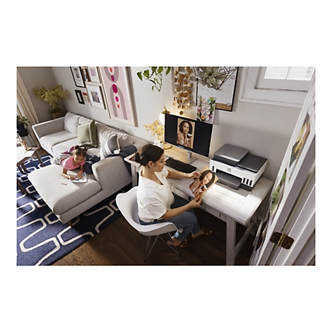 Une femme est assise à un bureau, regardant une photo d'elle-même sur un écran d'ordinateur. Un enfant dessine sur un canapé.