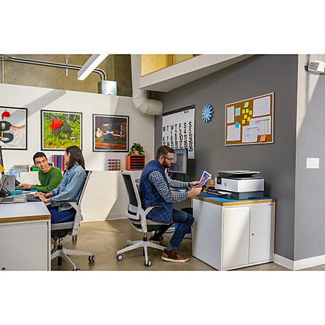 Büro mit drei Personen, Computern und einem Drucker. Eine Person sitzt vor dem Drucker.