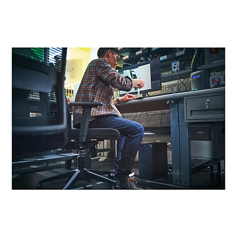Eine Person sitzt an einem Schreibtisch und arbeitet an einem Computer. Er trÃ¤gt ein kariertes Hemd und Jeans. Der Hintergrund zeigt technische GerÃ¤te.