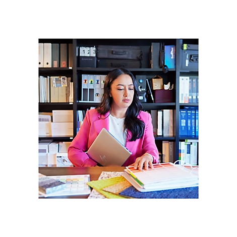 Frau in pinkem Blazer sitzt an einem Tisch, Laptop und Farbmuster vor sich. BÃ¼cherregal im Hintergrund.