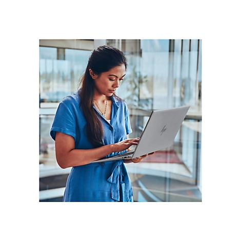Eine Frau in blauem Kleid arbeitet an einem Laptop. Sie steht vor einem Fenster mit Blick auf eine urbane Umgebung.