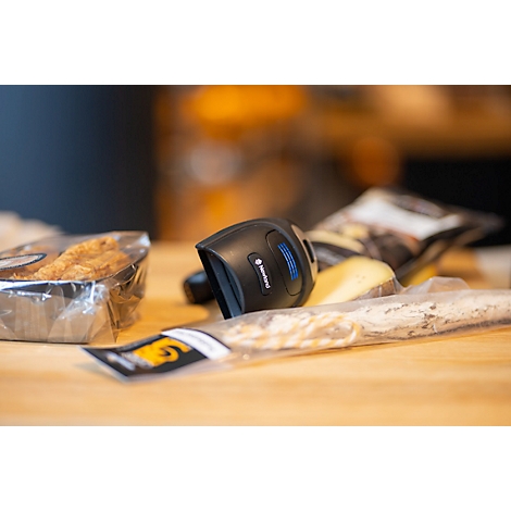 Nahaufnahme eines Barcodescanners auf einem Holztisch, umgeben von verpackten Lebensmitteln wie Käse und Gebäck.