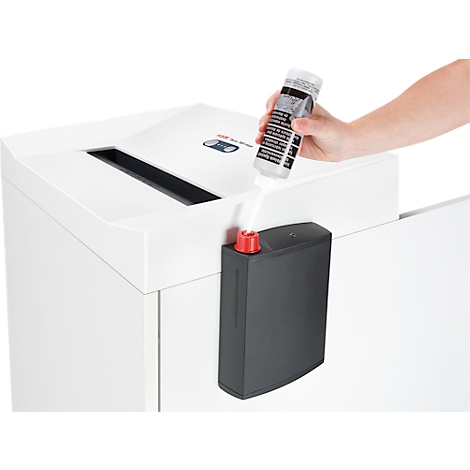 A hand pours liquid from a bottle into a paper shredder, presumably lubricating oil. Black tank with red cap below.