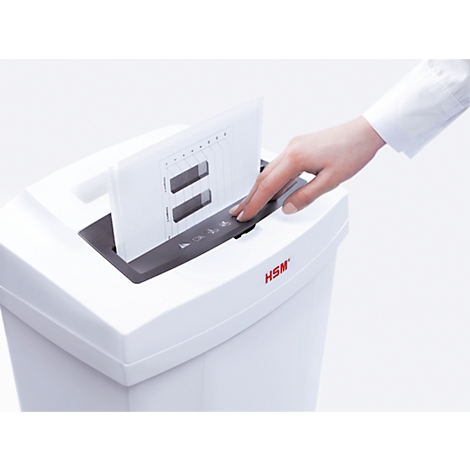 A person places paper into a shredder. The paper has lines and rectangular holes. The shredder is white with the inscription "HSM".